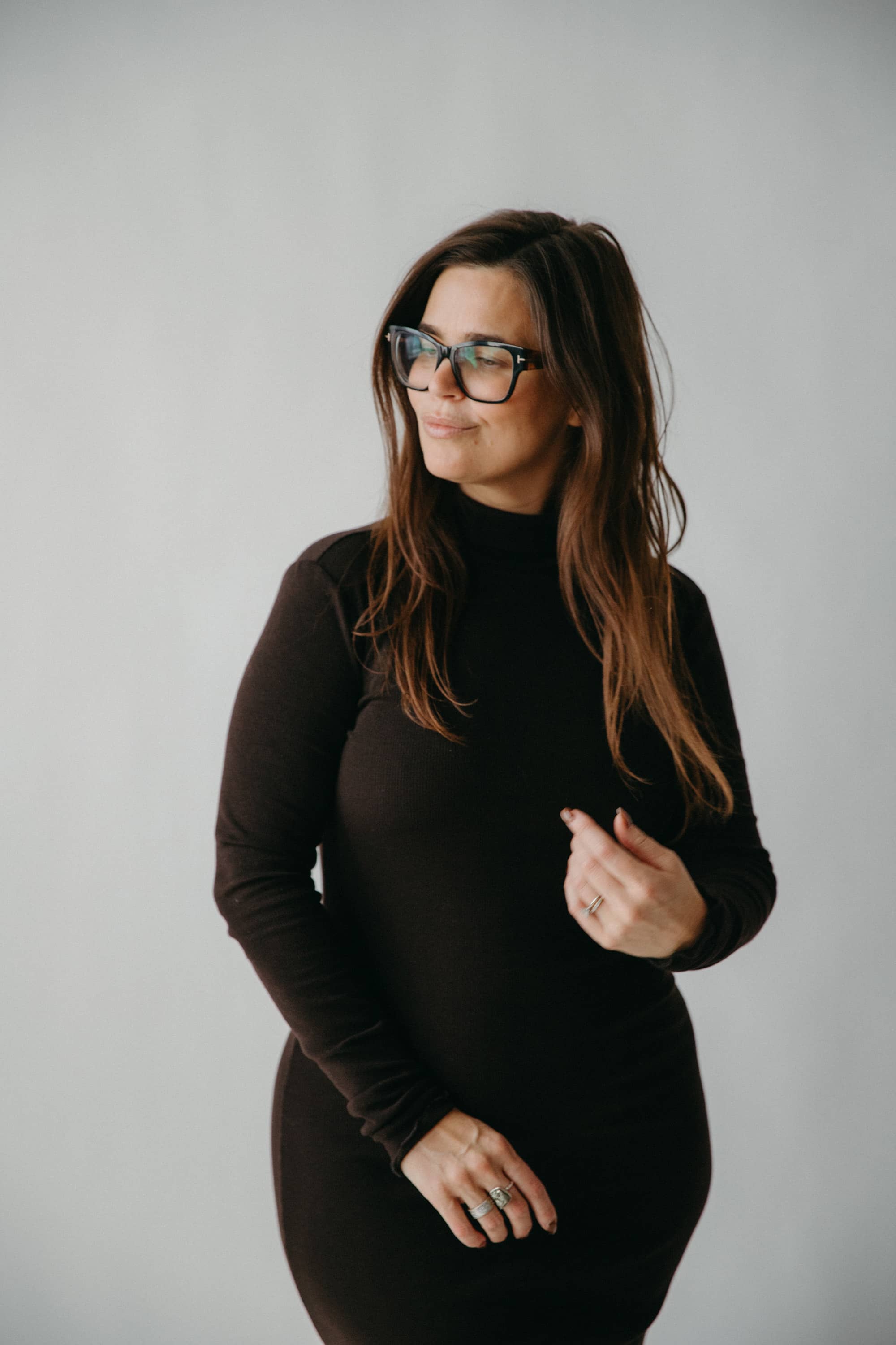 Woman wearing a black turtleneck dress and glasses against a plain background.