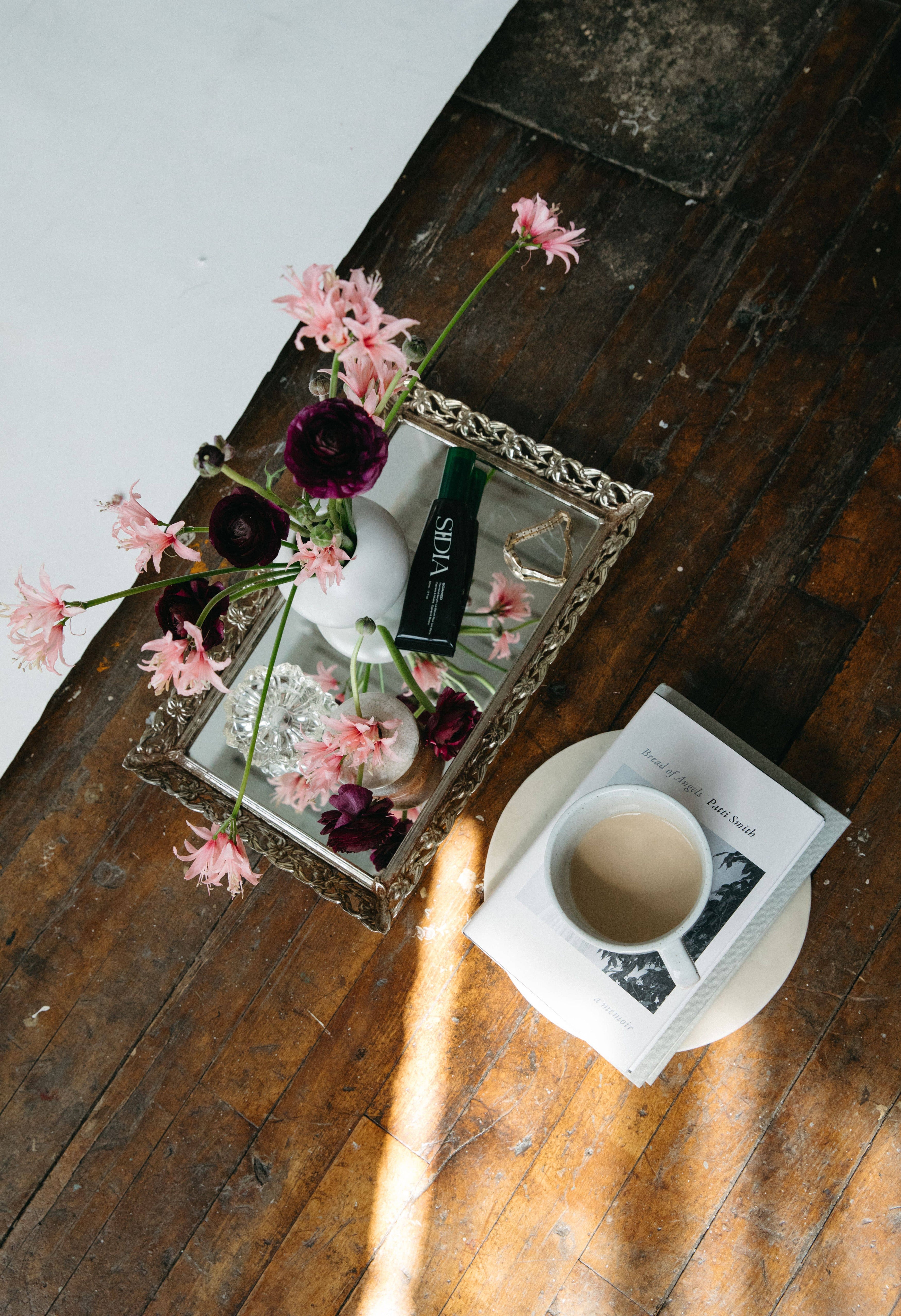 Tray with flowers and trinkets next to a coffee cup on a book, both on a wooden surface.